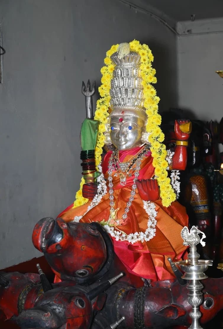 A silver deity figure in a tall crown, garlanded with yellow and white flowers, seated on a red wooden Nandi, with painted wooden figures behind and a silver oil lamp alongside